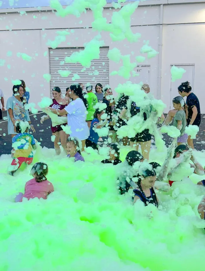 Teens, adults and kids play in bright green foam on a paved lot beside a building