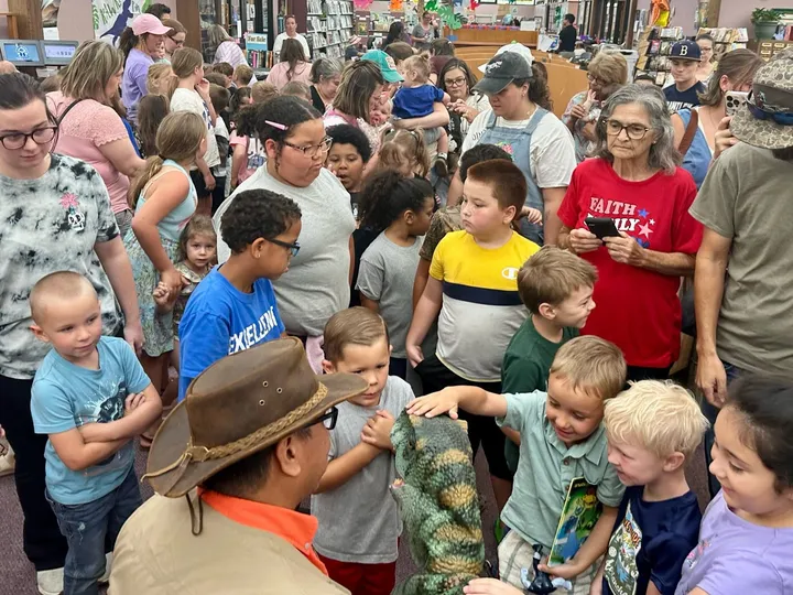 Families crowd a library floor around a Dinosaur Trainer and a baby dinosaur
