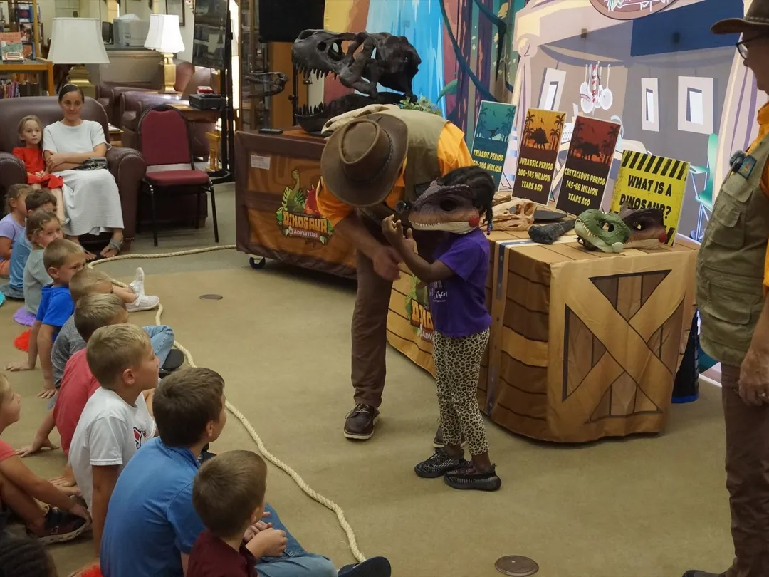 Children seated on the floor watch a dinosaur program as a young guest in a dinosaur mask meets the host