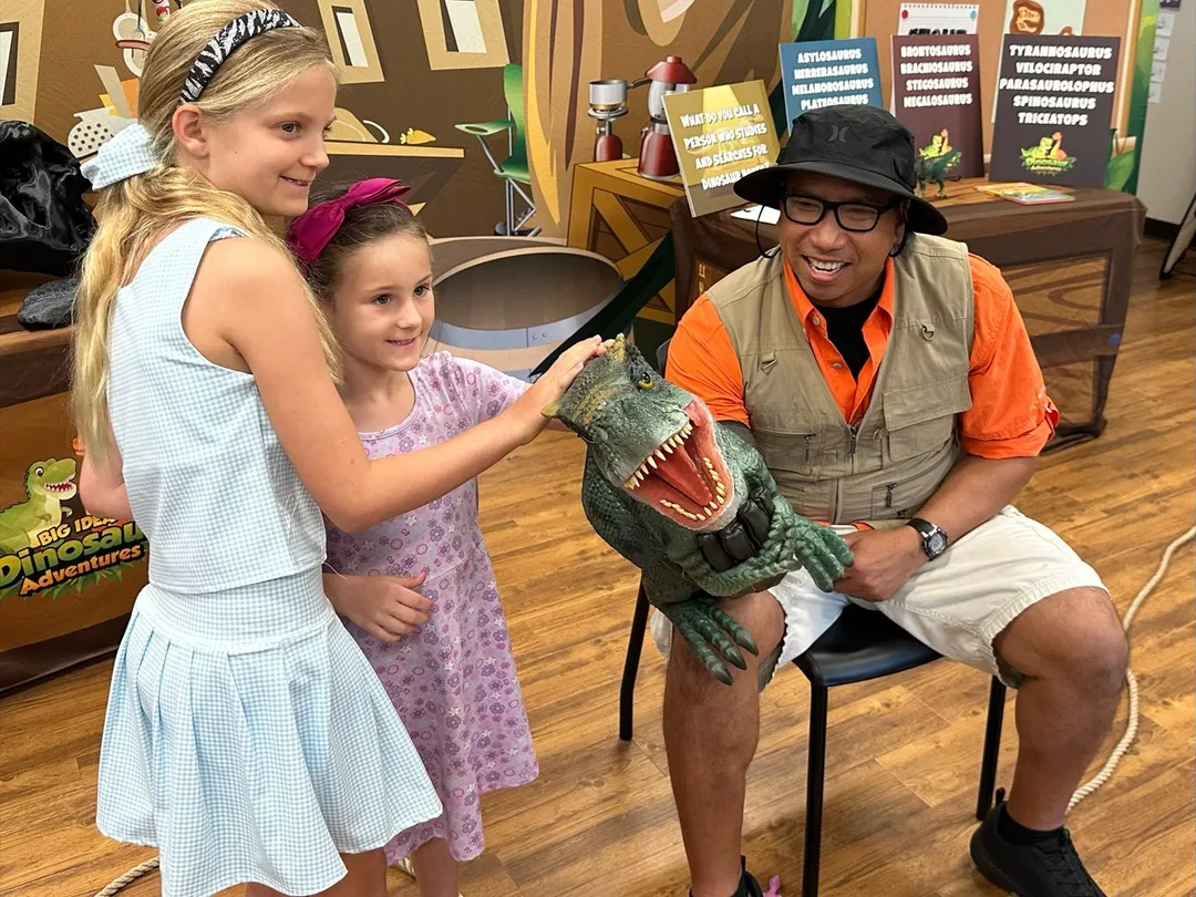 Two girls pet a roaring T. rex puppet during a dinosaur program