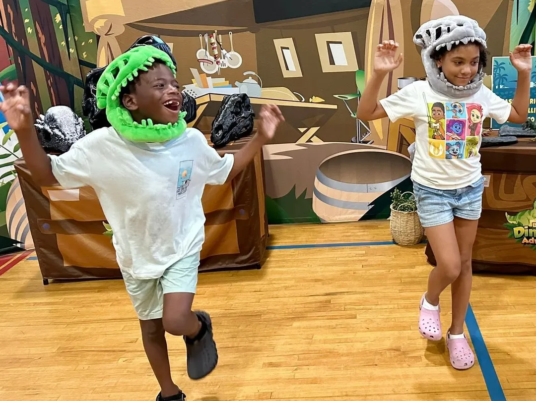 Two kids in plush dinosaur hats dance during an indoor dinosaur program