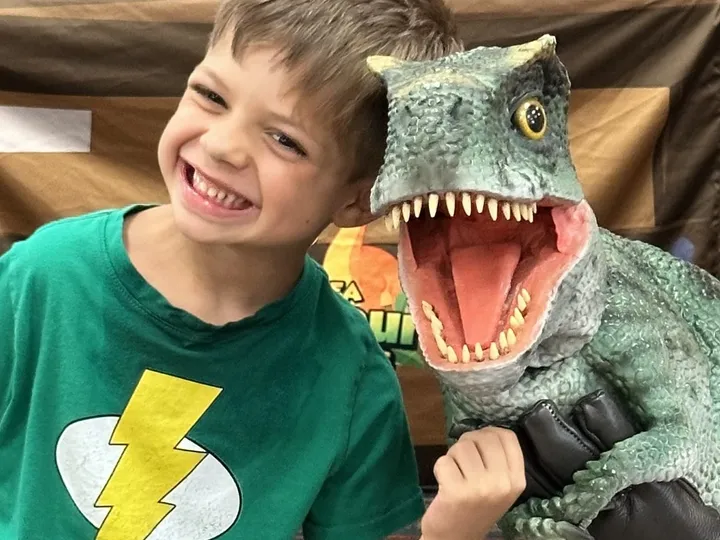A child grins beside a dinosaur puppet during an indoor dinosaur program