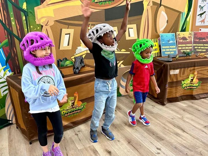 Kids in dinosaur skull hats cheer in front of a dig-site backdrop