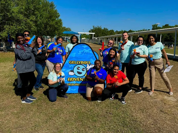 A team of adults poses with gel blasters beside a game banner on a sunny field