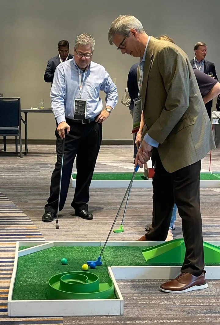 Two professionals in business attire take turns on a portable mini golf hole at a conference